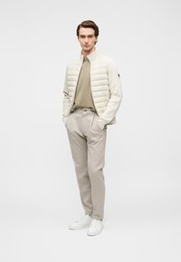 Leichte Steppjacke in Creme, mit Kragen und Reißverschluss. Darunter olivgrünes T-Shirt, kombiniert mit beigen gestreiften Hosen und weißen Sneakern.