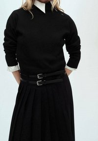 Personne portant un pull noir par-dessus une chemise blanche, assorti à une jupe plissée noire et une ceinture noire à double boucle.