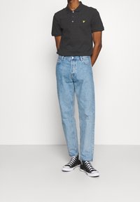 Černé polo tričko se žlutým logem v kombinaci se světle modrými džíny rovného střihu a černými vysokými teniskami. Minimalistický design s klasickým střihem.