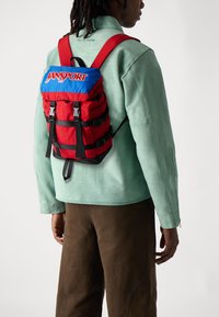 Sac à dos JanSport rouge et bleu avec des bretelles réglables, des fermetures à boucles et un design compact. Fabriqué en tissu avec une finition texturée.