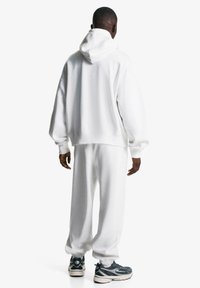 Homme portant un hoodie blanc et des survêtements, se tenant de dos, avec des baskets sombres sur un fond blanc uni.
