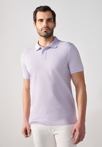 Polo shirt lavandă deschisă, realizat dintr-un material neted, cu guler clasic, mâneci scurte și o siluetă ajustată, asociat cu pantaloni albi.