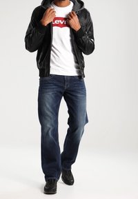 Svart läder bomberjacka med huva, vit t-shirt med röd 'Levi's' logotyp, blå raka jeans och svarta sneakers.