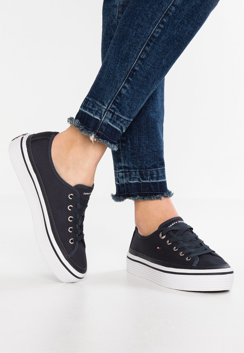 Pieds portant des baskets basses en toile noire avec des semelles blanches épaisses et un jean bleu foncé à ourlet effiloché, debout sur un sol blanc.