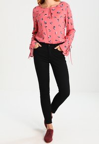 Blus i rosa med ett geometriskt mönster, kombinerad med svarta skinny jeans. Blusen har en rund hals och knyt detaljer vid ärmarna.