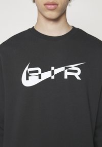 Personne portant un sweat à capuche noir avec un logo "AIR" stylisé blanc et un motif de swoosh sur la poitrine.