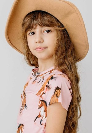 Young girl with long brown hair wearing a tan wide-brim hat and a pink shirt with horse illustrations, looking calmly at the camera.