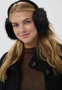 Čierne umelé kožušinové náušníky, okrúhle a nadýchané, nosené cez béžový sveter a čierny kabát s textúrovaným povrchom a veľkými čiernymi gombíkmi.