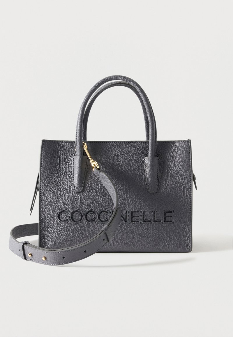 Sac à main en cuir gris texturé avec poignées supérieures, bandoulière amovible et nom de la marque embossé "COCCINELLE" sur le devant.