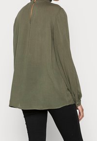 Blusa verde oliva a maniche lunghe con colletto alto e apertura a chiave sul retro. Presenta polsini con bottoni e un tessuto liscio e fluente.