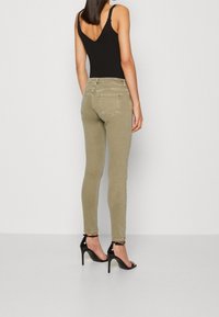 Jeans skinny verdes oliva con un diseño de yoke en la parte trasera, dos bolsillos traseros y una textura suave, combinados con sandalias de tacón negro.