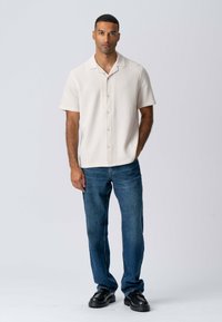 Kortærmet cremet skjorte med knapper og et struktureret mønster, parret med blå lige jeans og sorte sko. Enkel, moderne design.