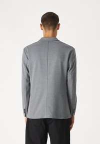 Grå blazer med texturerat tyg, enkel slits, spetsiga revers och detalj med tre knappar vid ärmslutet, sedd bakifrån mot en vit bakgrund.