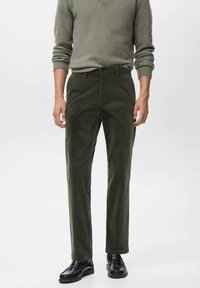 Pantalones de pana en verde oscuro, con corte recto, bolsillos laterales y cierre de botón. Llevados con un suéter gris y zapatos negros.