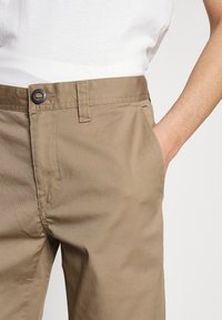 Beige puuvillachinohousut, joissa on tasainen etuosa, yksi takatasku ja metallinen nappisulku, sekä sileä pinta ja suora lahkeiden muoto.