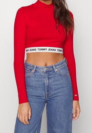 Kobieta w czerwonej długorękawowej krótkiej bluzce z napisem "Tommy Jeans" na pasku oraz w niebieskich, wysokich spodniach dżinsowych, stojąca na jednolitym tle.