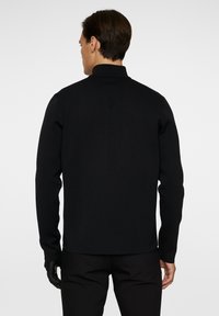 Svart stickad tröja med hög krage, strukturerat mönster och långa ärmar. Baksidan visar subtil märkning längs ryggraden.