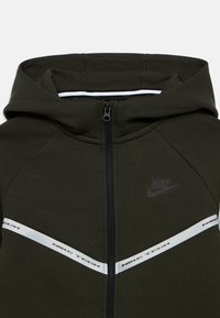 Σκούρα πράσινη ζακέτα με φερμουάρ διαθέτει δομημένο κουκούλι, λευκές λεπτομέρειες και λογότυπο "Nike Tech", κατασκευασμένη από μαλακό, υφή υλικό.