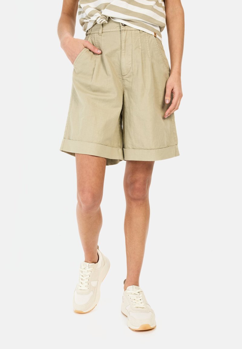 Helle khakifarbene Shorts mit hoher Taille, Falten, umgeschlagenen Säumen und seitlichen Taschen. Hergestellt aus leichtem Stoff. Kombiniert mit weißen Sneakers.