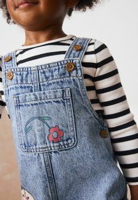 Denim overalls fából készült gombokkal és virágmintával, fekete-fehér csíkos hosszú ujjú pólóval párosítva. Világoskék szín.