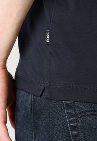 Närbild av en person som bär en mörkblå t-shirt med BOSS-logotyp och blå jeans, där sidofickans söm och fållens detaljer visas.