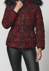 Czerwona i czarna pikowana kurtka zimowa z czarnym futrzanym wykończeniem przy kapture, z kieszeniami na zamek, zestawiona z czarnymi skórzanymi spodniami.