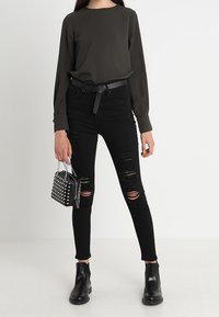 Jeans skinny negros desgastados, blusa de manga larga verde oscura, cinturón negro y botines. Pequeño bolso negro con tachuelas plateadas.