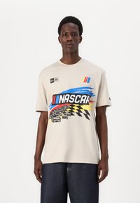 T-shirt en coton beige à manches courtes avec un motif NASCAR en bleu, rouge et jaune, ainsi que des accents de motif à damier et de logo.