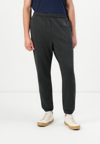 Persona vestida con pantalones joggers elásticos de cintura en gris oscuro con los tobillos ajustados y zapatillas blancas con cordones, de pie frente a un fondo liso.