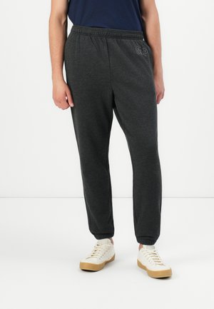 Person, der eine dunkelgraue Jogginghose mit elastischem Bund und Bündchen an den Knöcheln sowie weiße Schnürschuhe trägt, steht vor einem einfarbigen Hintergrund.