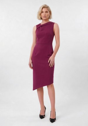 Γυναίκα που φοράει αμάνικο, ασύμμετρο, σκούρο μοβ φόρεμα μήκους μέχρι το γόνατο με μαύρες γόβες, στέκεται μπροστά σε απλό φωτεινό φόντο.