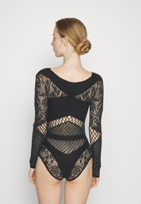 Musta bodysuit, jossa on pitkät hihat, pitsi- ja kalaverkkomallit, leikattu yksityiskohta sekä kiiltäviä yksityiskohtia koko designissa.