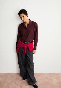 Burgundy long-sleeve button-up shirt paired with wide black trousers, accented by a red sweater tied around the waist and black sleek shoes.