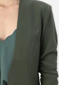 Donkergroene blazer met een gladde textuur, ontwerp met één knoop en een getailleerde pasvorm. Daaronder bevindt zich een gelaagd groene top met een geschulpte halslijn.