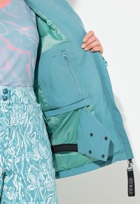 Tyrkysová bunda se zapínací kapsou, podšívkou a kalhotami s květinovým vzorem. Obsahuje patentky a zipový detail na spodním lemu.