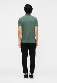 Polo verde de mangas curtas, tecido texturizado, com um design nas costas apresentando detalhes em linhas horizontais. Calças pretas e sapatos completam o visual.