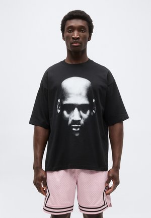 Bărbat purtând un tricou negru supradimensionat cu o imprimare mare în tonuri de gri a unui chip și pantaloni scurți de baschet roz cu margini negre, stând pe un fundal simplu și luminos.