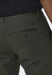 Groene broek met een visgraatpatroon, voorzien van een achterzak met knoopsluiting. De stof heeft een gestructureerd uiterlijk.