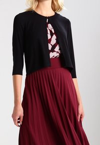 Black cropped cardigan with 3/4 sleeves, button closure at the neck; worn over a burgundy pleated dress with a patterned bodice.