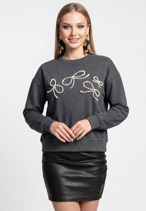 Ung kvinde iført mørkegrå sweatshirt med perle-sløjfe-designs og sort lædernederdel, smilende med hænderne foldet foran.