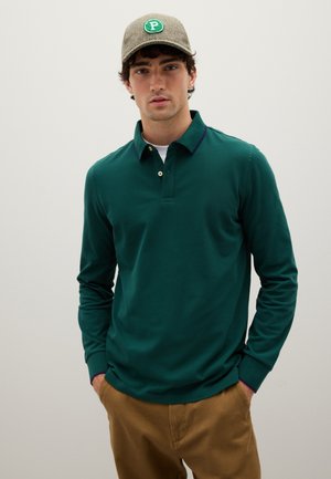 Hombre vistiendo una camiseta polo de manga larga verde, pantalones marrones y una gorra de cuadros con un parche verde de "P", de pie contra un fondo claro liso.