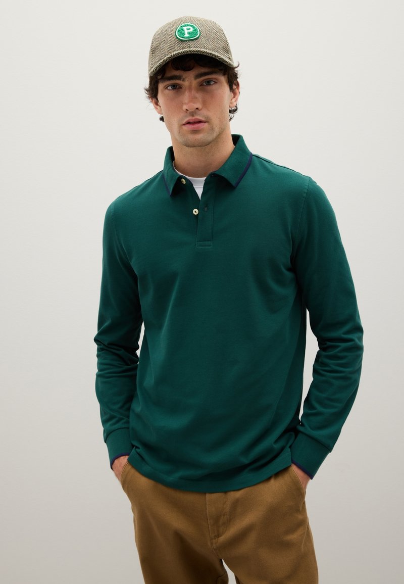 Mann trägt ein grünes Langarm-Polohemd, braune Hose und eine karierte Mütze mit einem grünen "P"-Patch, im Stehen vor einem schlichten hellen Hintergrund.