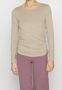 Beige long-sleeve top with a round neck and side ruching details. Worn with lavender wide-leg pants. Smooth texture, fitted design.