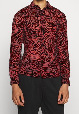 Camicia rossa con bottoni e strisce zebrate nere indossata con pantaloni neri, mostrata dal collo fino alla parte superiore delle cosce su uno sfondo semplice.