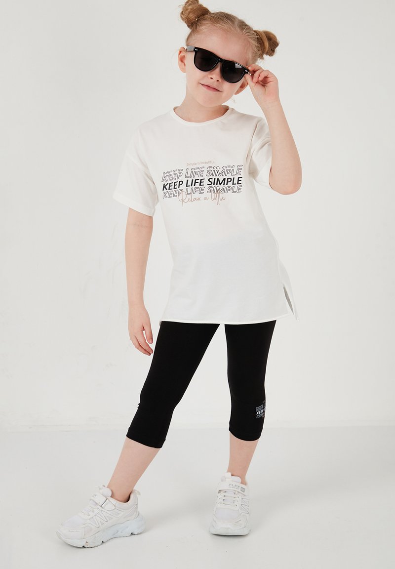Jeune fille portant des lunettes de soleil, un t-shirt blanc "Keep Life Simple", des leggings courts noirs et des baskets blanches, se tenant devant un fond uni.