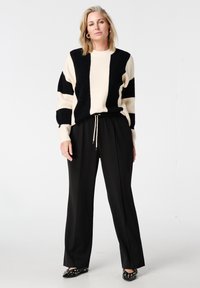 Pull rayé noir et crème à coupe décontractée, associé à un pantalon noir ajusté. La tenue comporte une taille à cordon de serrage et des accents décoratifs.