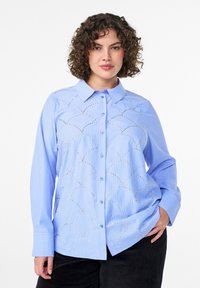 Light blue striped button-up shirt with long sleeves, featuring decorative scalloped stitching and metallic accents on the front.