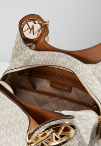 Sac à main en toile beige avec un motif monogrammé, accents en cuir marron, ferrures dorées et un intérieur spacieux comprenant plusieurs compartiments.