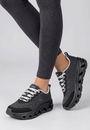 Personne portant des leggings gris foncé et des chaussures de sport noires et grises avec des semelles épaisses et des lacets blancs, debout sur une surface claire.