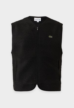 Homme portant un gilet noir sans manches sur un polo gris, pantalon bleu marine et baskets blanches, debout devant un fond uni.
